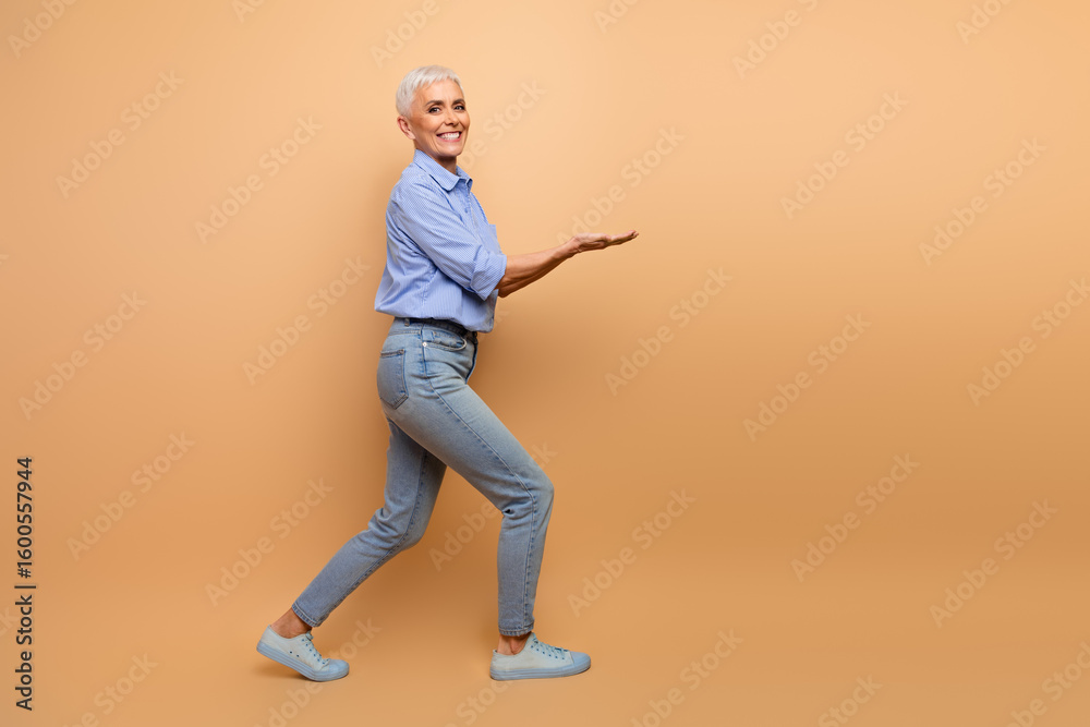 Elegant smiling mature woman presenting with hand gestures against a beige background, showcasing casual style in blue striped shirt. Elegant smiling mature woman presenting with hand gestures against a beige background, showcasing casual style in blue striped shirt.
