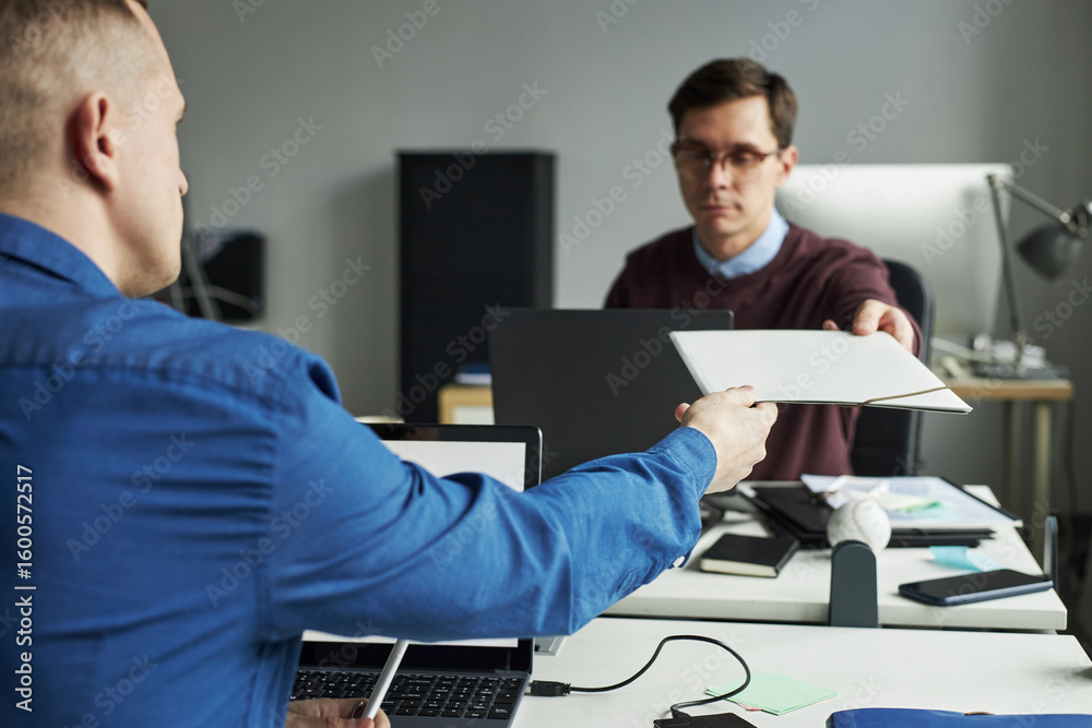 Fototapeta premium Caucasian man handing document folder to another male colleague sitting at desk in modern office, both focused on exchanging paperwork during business meeting. collaborating on presentation