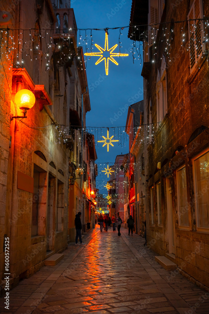 Fototapeta premium Beautiful Christmas decorations with star-shaped lights and glowing garlands adorn a cobbled pedestrian street in Poreč’s historic center