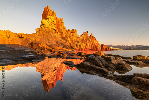 View of rugged red rocks reflecting in calm waters under a bright sky, creating a striking contrast of color and texture, Arbatax, Sardegna, Italy.