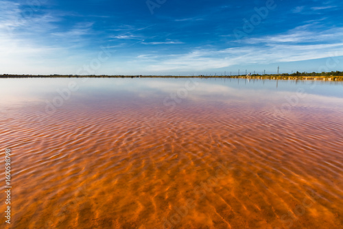 View of a vibrant pink salt lake reflecting the clear blue sky and wispy clouds, creating a surreal and colorful landscape, Saline di Cervia, Emilia-Romagna, Italy.