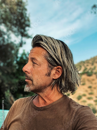 Tanned man with blond half long hair and grey stubble beard in brown t-shirt enjoys sunny summer weather in garden in Spain.