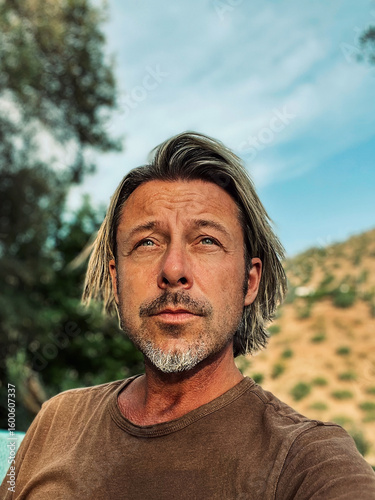 Tanned man with blond half long hair and grey stubble beard in brown t-shirt enjoys sunny summer weather in garden in Spain.