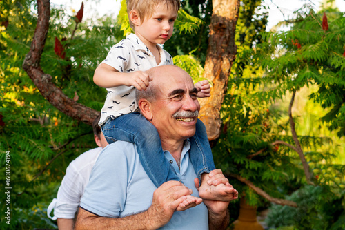 Joyful grandfather smiles warmly while carrying his young grandson on his shoulders, sharing a playful and affectionate moment outdoors in a lush summer garden