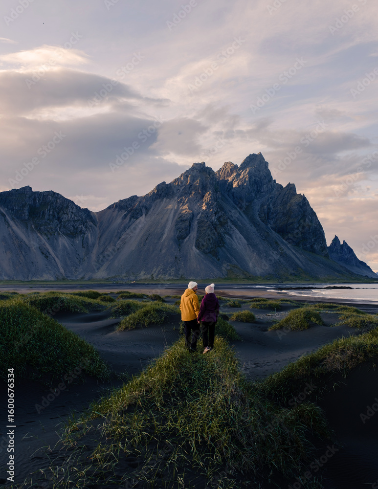 Fototapeta premium Two travelers explore lush grass and black sand dunes beneath Vestrahorn mountain in Stokksnes, Iceland, embodying adventure in a stunning twilight landscape.