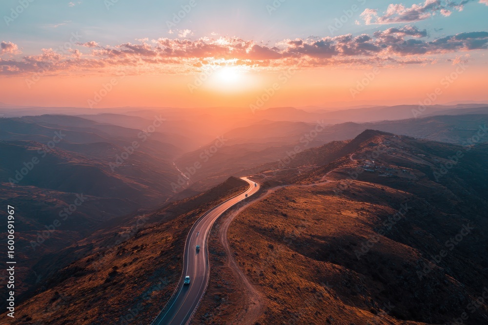 Fototapeta premium Winding road across mountaintop with vehicles during orange sunset over landscape