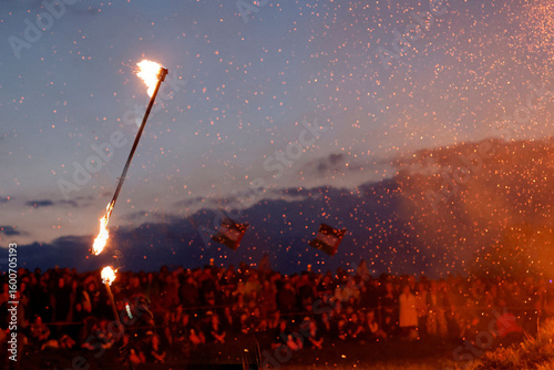 Fiery spectacle at night with two flaming torches held high above a large crowd, sparks raining down.