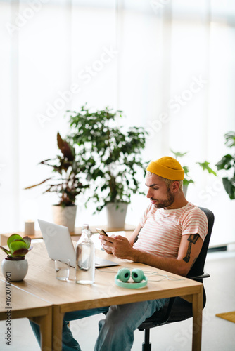 A young man in a yellow beanie and striped shirt sits at a desk, focused on his smartphone. A laptop, water bottle, and headphones are nearby. Plants decorate the bright background.