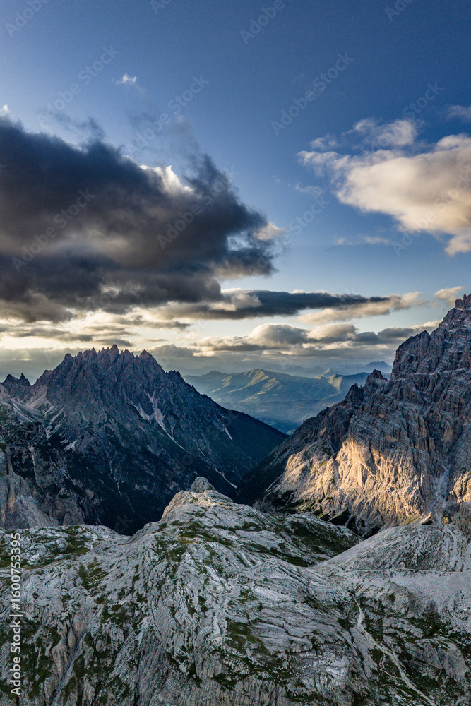 Obraz premium Rugged peaks and dramatic valleys in the Dolomites at sunset — vertical aerial view with scattered light on alpine ridges, dark forests below, and heavy clouds above the Italian Alps