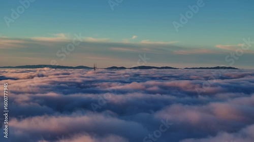Aerial view of Sutro Tower poking through a sea of fluffy clouds, with mountains in the distance under a blue sky, San Francisco, California, United States.