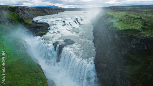 Fotografie Gullfoss waterfall pours dramatically over cliffs into a fierce river below, surrounded by lush green landscapes