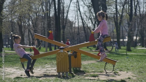 Little girls swing on the see-saw at the playground in the park, with trees in the blurred background, during the spring season.