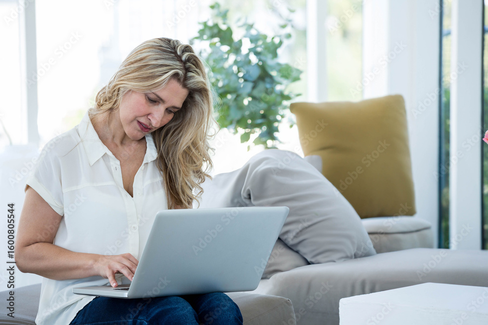 Woman typing on silver laptop while sitting on light-colored sofa under floor-to-ceiling windows Woman typing on silver laptop while sitting on light-colored sofa under floor-to-ceiling windows