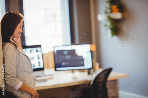 professional leaning against wall near window at office holding smartphone by desk with monitors