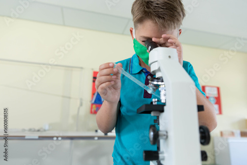 Boy wearing green mask peering into white microscope holding pipette and slide at classroom bench