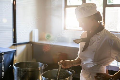 Chef wearing white coat and hat stirring pot on stovetop as sunlight streaming through windows