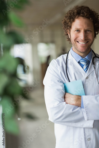 Male doctor standing in hospital hallway holding blue folder and wearing stethoscope, copy space