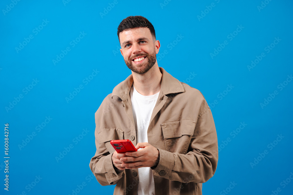 Portrait of a cheerful young bearded man in a beige jacket and whit shirt, holding a red smartphone and smiling confidently against a solid blue studio backdrop, symbolizing modern digital Portrait of a cheerful young bearded man in a beige jacket and whit shirt, holding a red smartphone and smiling confidently against a solid blue studio backdrop, symbolizing modern digital