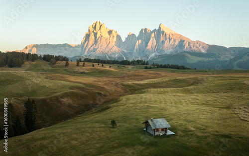 Aerial view of a rustic cabin nestles in a valley as the sun bathes the Sassolungo and Sassopiatto peaks in warm hues, Via Alpe di Siusi, Busto Arsizio, Lombardia, Italy.