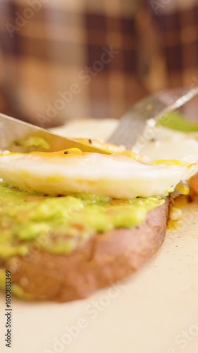 Vertical video. Close up of fork and knife cutting through runny yolk egg on avocado toast with salad on side in warm indoor setting, Healthy Eating Concept