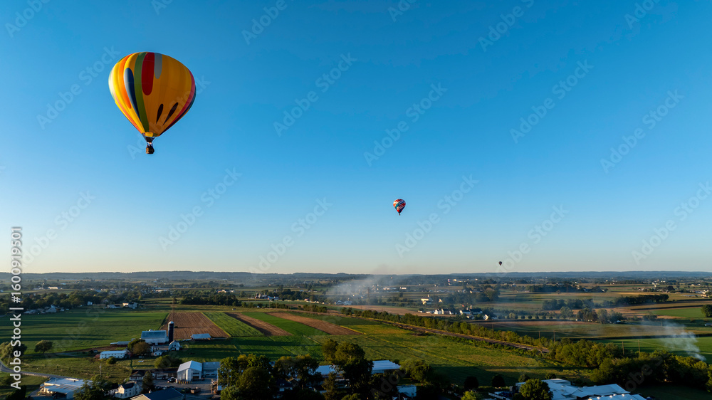 Obraz premium Hot air balloons gracefully rise against a clear blue sky, gliding over expansive green fields and farms during the early morning hours. The tranquil landscape showcases the beauty of nature.