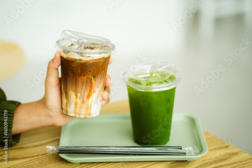 Tablou pe pânză hand of woman holding a take-out iced coffee with iced matcha green tea on wooden table at cafe