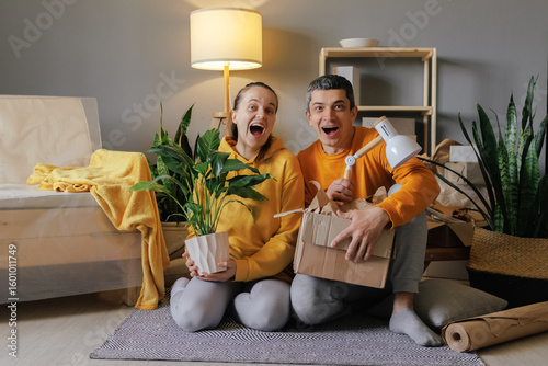 Fényképezés A delighted couple sits on the floor of their new home, holding a houseplant and a box of items, looking surprised and excited about their move