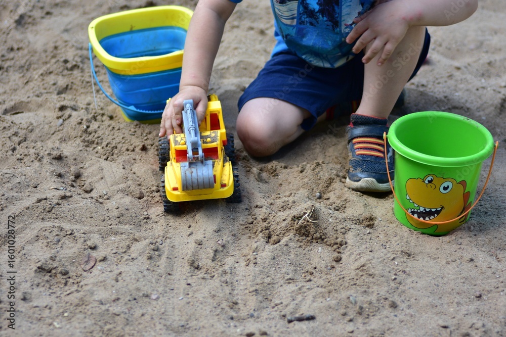 Anonymous toddler boy playing in the sandbox with a yellow toy truck and two buckets, surrounded by dry sand on a sunny day, captured during early childhood play activity. Anonymous toddler boy playing in the sandbox with a yellow toy truck and two buckets, surrounded by dry sand on a sunny day, captured during early childhood play activity.