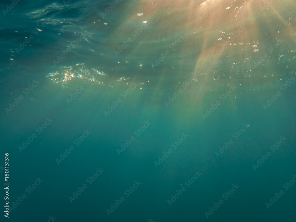 Fototapeta premium Dark blue ocean surface seen from underwater. Abstract waves underwater and rays of sunlight shining through, Sun light rays undersea deep, Underwater background with sea bottom, Mediterranean sea.