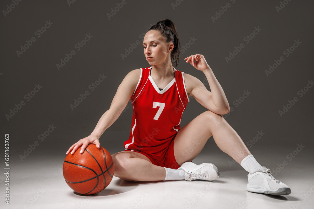 Naklejka premium Female basketball player in red uniform sits on floor with ball, athletic woman, studio portrait