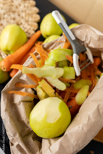 Paper bag with organic waste from apples and carrots next to a vegetable peeler. Organic waste, sorting.