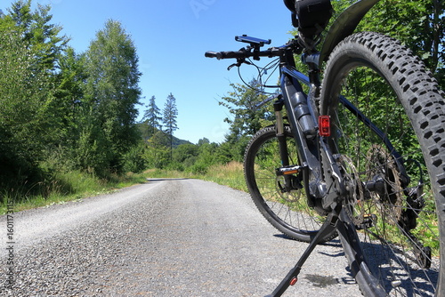 A mountain bike with thick tires, parked on a gravel road. The road stretches into the distance, surrounded by green trees. A clear blue sky. Outdoor setting for cycling.