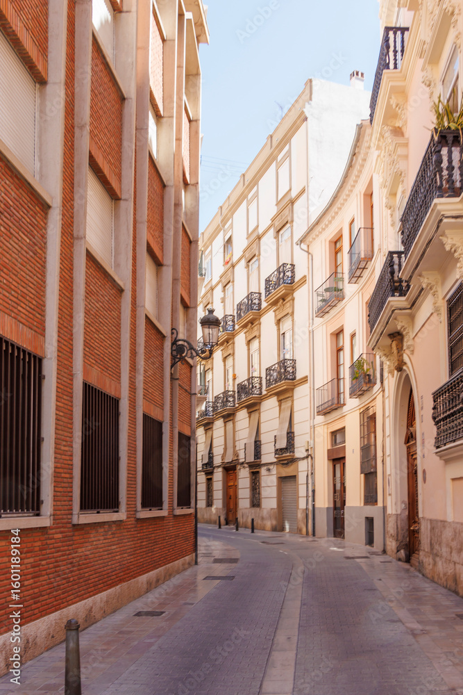 Naklejka premium Charming narrow street in Valencia featuring traditional Spanish architecture, with decorative balconies and sunlight illuminating the vibrant buildings, creating a serene urban atmosphere.