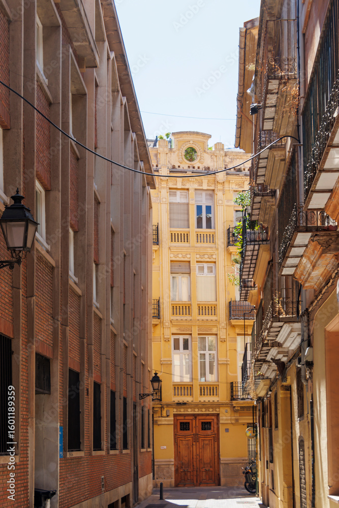Fototapeta premium Scenic view of a narrow street in Valencia, showcasing beautiful architecture, sunlight, and contrasting building styles capturing the essence of urban life.