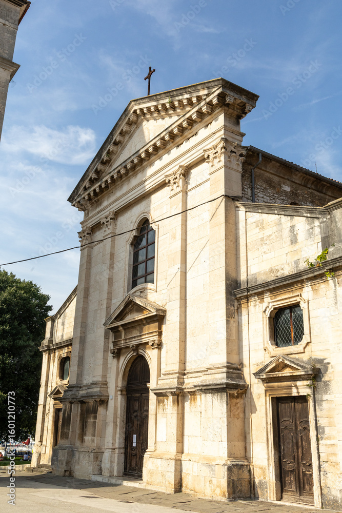 Fototapeta premium Pula, Croatia - July 15, 2025: Historic stone church with intricate architecture and cross on top, surrounded by trees and blue sky