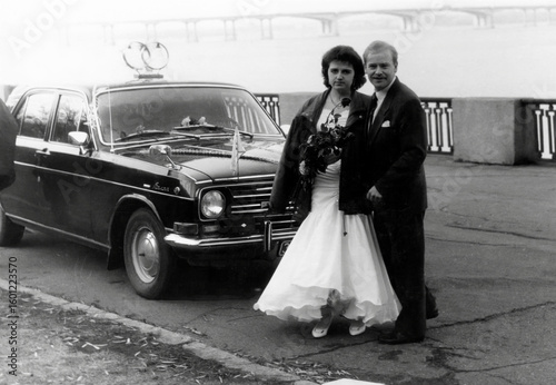 Vintage portrait of bride and groom on their wedding day near the car on the river bank. Retro photo from 1992.