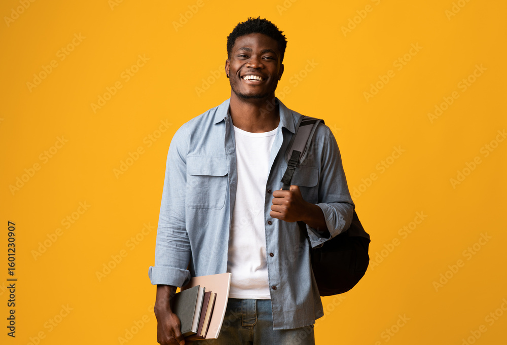 Fototapeta premium Positive millennial black man student in casual posing with books and notepads on yellow studio background, young african american guy enjoying studying at university or college, copy space