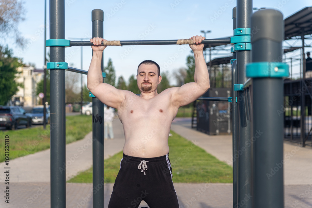 Fototapeta premium Muscular man doing pull-ups on outdoor gym equipment. Shirtless, he showcases defined upper body strength. Set in a sunny park, the photo captures a healthy lifestyle