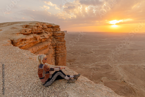 Female tourist sitting on the edge of the world escarpment at sunset, contemplating the cliffs of Tuwaiq mountain range in Riyadh, Saudi Arabia, a breathtaking natural wonder