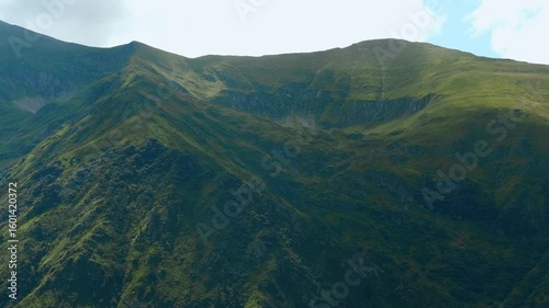 Mountain ridge covered with green grass. Sunlight illuminating lush green grass covers a mountain ridge. The camera pans gracefully along the ridge, showcasing the vibrant landscape under a blue sky