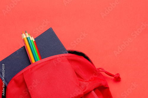 A red backpack with a notebook and colorful pencils on red background. Back to school concept with copy space.