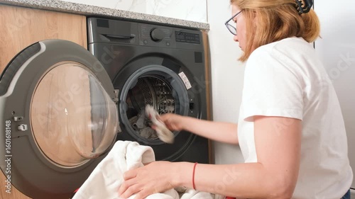 Young woman loading laundry into a front-loading washing machine at home. She kneels by a red basket full of white clothes in a modern laundry room