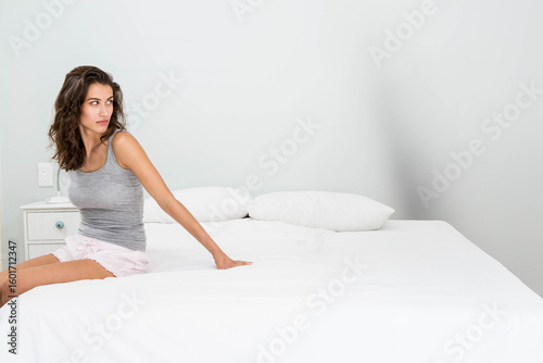 Woman sitting on bed edge near white nightstand with lamp, outlet and pillows in grey bedroom