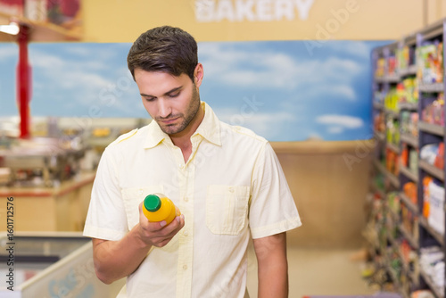 Male shopper standing in supermarket bakery aisle holding orange juice bottle and reading label