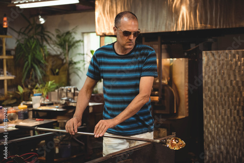 Middle-aged man standing at glassblowing bench gripping blowpipe with molten glass at furnace