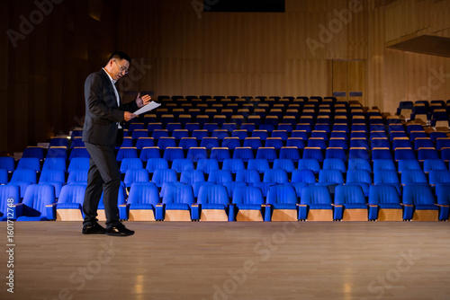 Εκτύπωση καμβά Asian mature man presenting on stage floor beside blue seats in hall holding pap