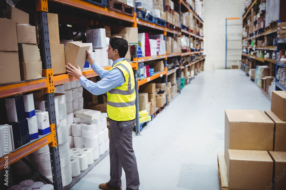 Fototapeta premium Warehouse worker reaching for cardboard box on tall metal shelf in brightly lit warehouse aisle