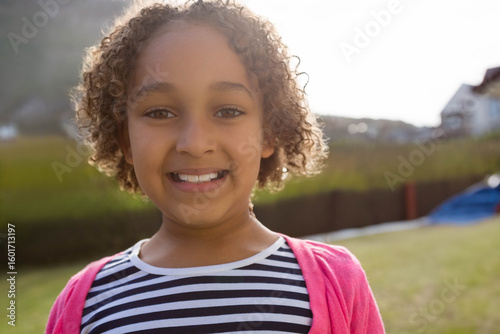 Child girl standing in grassy yard wearing striped shirt and pink cardigan under bright sky