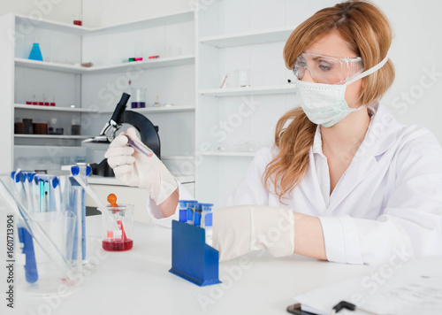 Pipette transferring red liquid into beaker on laboratory bench with test tube rack and microscope