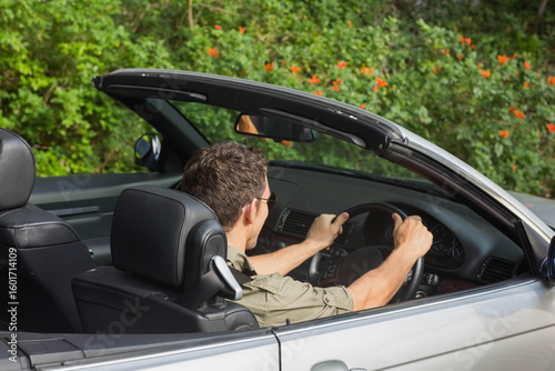 Silver convertible showing steering wheel on roadside against hedge dotted with orange flowers